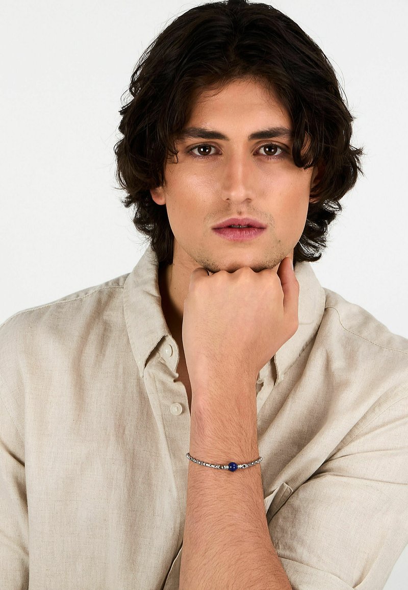 Bracciale argento con perla blu, indossato al polso. Modello vestito con una camicia di lino leggera e di colore neutro. Primo piano sui dettagli del bracciale.