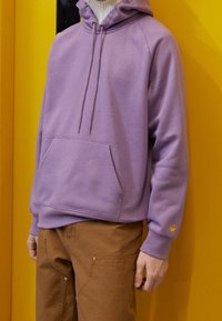 Person som bär en ljuslila hoodie med framficka och bruna byxor, stående mot en klar gul bakgrund.