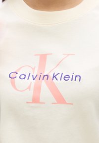 Ελαφριά μπεζ μπλούζα με στρογγυλή λαιμόκοψη, με ροζ και μωβ κεντημένο λογότυπο "Calvin Klein" και ένα στιλιζαρισμένο σχέδιο "CK".