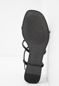Sandale en cuir noir avec une semelle plate et une texture antidérapante. Elle dispose de plusieurs brides tressées et d'éléments de design minimalistes.