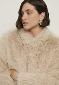 Beżowy płaszcz z futrzanym kołnierzem i długimi rękawami. Teksturowany, miękki materiał o luźnym kroju. Wyjątkowy detal w postaci srebrnego kolczyka.