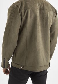 Olive green suede jacket with quilted shoulder accents, zippered cuffs, and a smooth texture. Features a button closure at the hem.