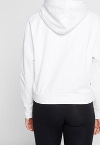 Felpa bianca con una texture morbida, caratterizzata da un cappuccio, spalle scese e un elastico in vita, abbinata a leggings neri.