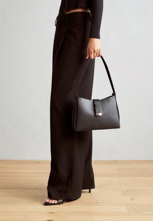 Black handbag with structured shape, smooth texture, and a metallic clasp. Paired with wide black pants and open-toe sandals on wooden floor.