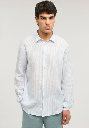 Junger Mann mit lockigem dunklem Haar, der ein weißes langärmeliges Hemd mit dezenten vertikalen blauen Streifen trägt und vor einem einfarbigen Hintergrund steht.