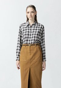 Blouse à carreaux noir et blanc à manches longues, associée à une jupe brune taille haute avec une fente à l'avant et des poches.