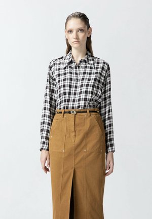 Blusa a cuadros en blanco y negro con mangas largas, combinada con una falda marrón de cintura alta que tiene una abertura frontal y bolsillos.
