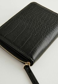 Portefeuille noir texturé avec un design en relief de crocodile, une fermeture éclair dorée et une sangle pour le poignet. Le matériau semble synthétique et a une finition lisse.