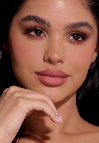 Close-up van een gezicht met een gladde huid, volle lippen in matte mauve, donker haar, gedefinieerde wenkbrauwen en subtiele oogmake-up. Hand rust nadenkend op de kin.