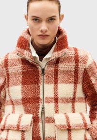 Plaid fleece jacket in cream and rust with a zip front, collared neckline, and two front pockets. Soft texture with a warm appearance.