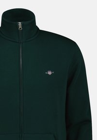 Dunkelgrüner Reißverschluss-Pullover mit hohem Kragen, die Stofftextur ist sichtbar, und es befindet sich ein kleines gesticktes Logo auf der linken Brust.