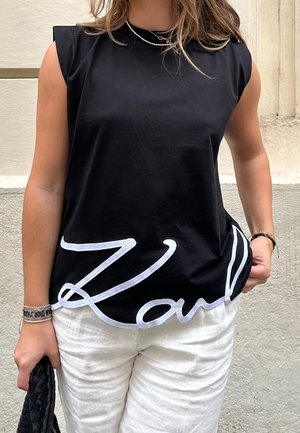 Femme portant un haut noir sans manches avec le texte en écriture cursive blanche "Karl" et un pantalon blanc, tenant un sac noir tissé, debout contre un mur texturé.
