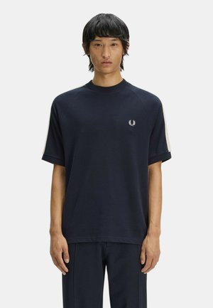 Giovane uomo con capelli neri indossa una camicia blu navy a maniche corte con strisce bianche sulle spalle e un piccolo logo bianco sul petto.