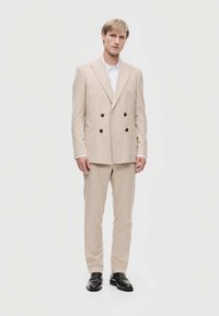 Beige Doppelreihiger Anzug mit zwei Fronttaschen und dunklen Knöpfen, kombiniert mit einem weißen Hemd und schwarzen Schuhen, schlanker Schnitt.