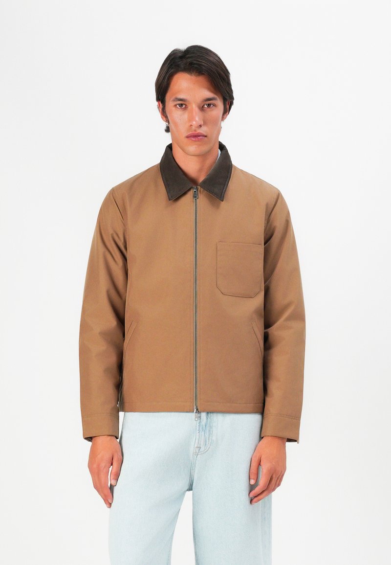 Man wearing a tan zip-up jacket with dark brown collar and light blue jeans, standing against a plain white background.