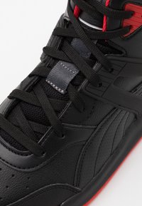 Schwarzer Sportschuh mit einem Obermaterial aus Leder, strukturierten Akzenten, roten und grauen Details sowie schwarzen Schnürsenkeln, mit auffälliger Naht an der Seite.