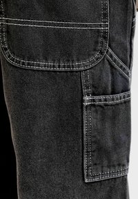 Σκούρο τζιν ύφασμα με εμφανή λευκή ραφή που περιγράφει δύο ορθογώνιες τσέπες στο πλάι του ρούχου.