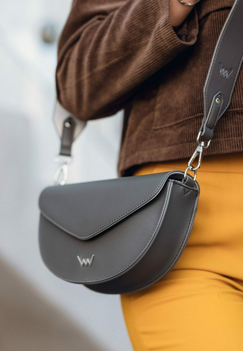 Šedá kožená crossbody taška s zakřiveným tvarem, klopovým uzávěrem a stříbrnými kovovými doplňky. Obsahuje logo a nastavitelný popruh.