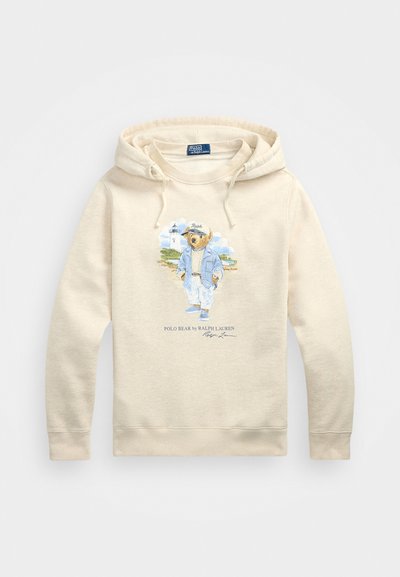 Beige kapucnis pulóver pamut keverékből, amelyen egy laza öltözetű medve grafikája látható. Zsinóros kapucnival és bordázott mandzsettákkal rendelkezik.