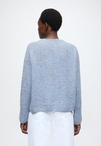 Ανοιχτό μπλε πουλόβερ με υφασμάτινη πλέξη και πολύχρωμες βούλες σε όλη την επιφάνεια. Μακριά μανίκια και στρογγυλεμένο τελείωμα, φορεμένο πάνω από λευκό παντελόνι.