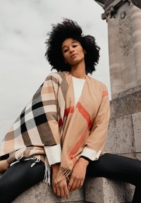 Châle beige à carreaux avec franges, aux accents rouges. Le modèle porte un pull en coton côtelé blanc et un legging noir, assis sur des marches en pierre.