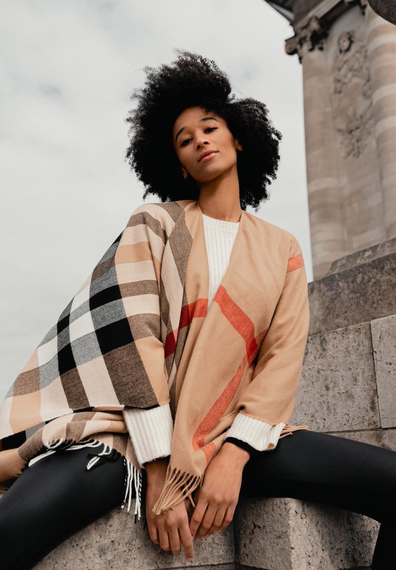 Châle beige à carreaux avec franges, aux accents rouges. Le modèle porte un pull en coton côtelé blanc et un legging noir, assis sur des marches en pierre.