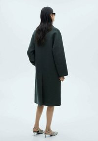 Lange, donkergroene wollen jas met verlaagde schouders en een rechte belijning. Gecombineerd met lichtgrijze schoenen met een slingback-ontwerp.