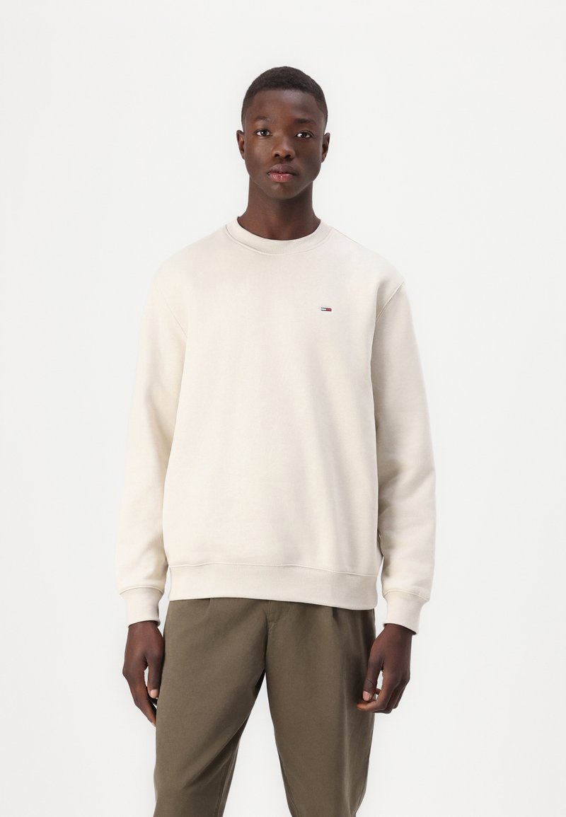 Lys beige genser med lange ermer, rund hals, ribbestrikkede mansjetter og kant. Liten logo detaljer på brystet. Båret med olivengrønne bukser.