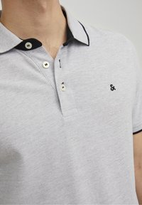 Ljusgrå pikétröja i en texturerad tyg, med kontrasterande marinblå collar och knappaccentuer, med en liten broderad logga på bröstet.