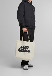 Osoba oblečená v černé mikině a kalhotách drží béžovou tašku s nápisem "GOOD KARMA!" v tučných černých písmenách, stoj�í na šedém pozadí.