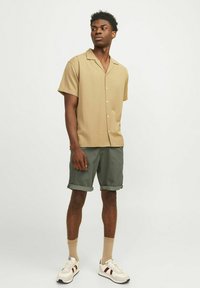 Chemise beige à manches courtes avec un col en V et des boutons, associée à des shorts roulés verts foncés et des baskets blanches avec des accents rouges.