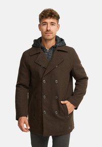 Cappotto doppiopetto marrone con bottoni neri, tasche laterali e una felpa grigia che spunta dal colletto; realizzato in tessuto texturizzato.