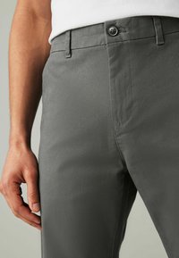 Pantaloni grigi realizzati in tessuto liscio, con zip e chiusura a bottone, e tasche laterali. Cuciture a vista lungo le cuciture.