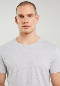 Ung mand med kort hår og blå øjne iført en ensfarvet lysegrå t-shirt mod en neutral baggrund.