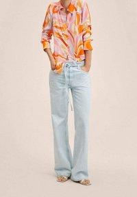 Camicia a maniche lunghe colorata con motivi a onde rosa, arancione e viola, abbinata a jeans a gamba larga azzurri chiari e sandali.