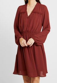 Vestido burdeos con mangas largas, escote en pico y detalle de encaje a lo largo del escote y el cuerpo; cintura elástica para un efecto fruncido.