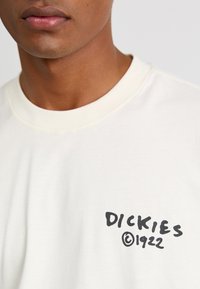Cream-colored cotton t-shirt with short sleeves; features a black graphic logo reading "DICKIES ©1922" on the left chest area.