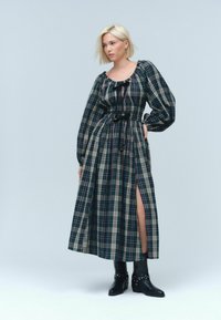 Lange geruite jurk met zwarte, groene en crème kleuren; gathered halslijn, lange mouwen, cinched taille en zijsplit. Gekleed met zwarte enkellaarzen.