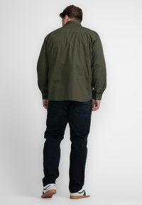 Camisa de botones de manga larga en verde oliva con patrón, combinada con jeans azul oscuro y zapatillas blancas, vista desde la espalda.