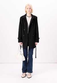 Un trench-coat noir à double boutonnage avec de grands boutons, associé à un chemisier blanc, un jean évasé bleu et un sac à main blanc. Des ballerines avec accents.