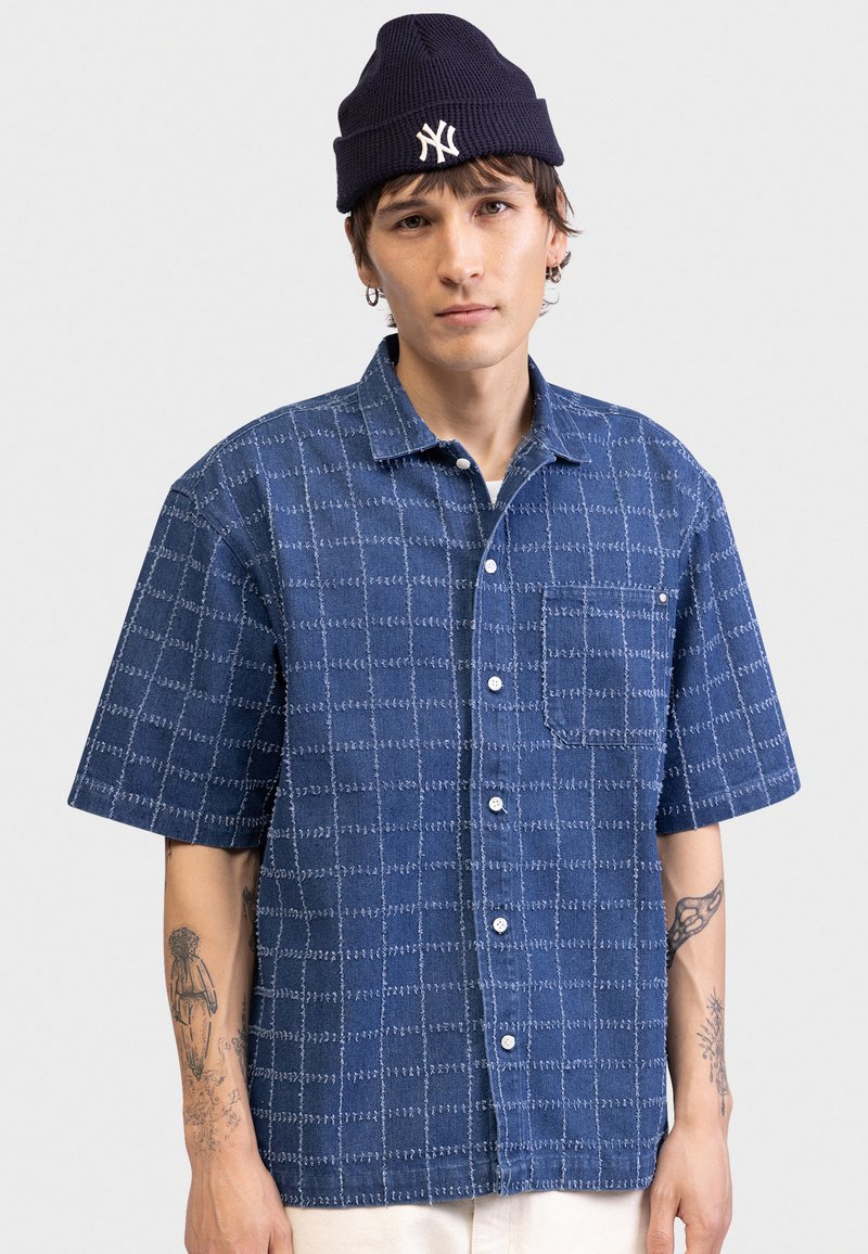 Νέος άνδρας που φοράει μπλε σκούφο με λευκό λογότυπο, μπλε πουκάμισο με κοντά μανίκια και καρό σχέδιο, και ορατά τατουάζ και στα δύο αντιβράχια.