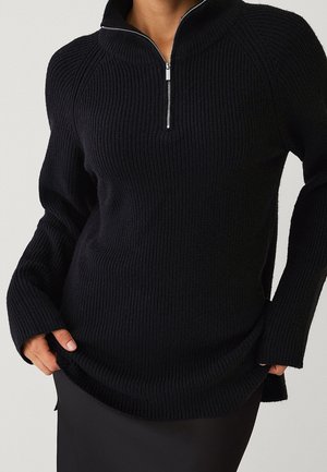 Henkilö pukeutunut mustaan ribbineulottuun neulepaitaan, jossa on puolivetoketjullinen kaulus, kädet hieman neuleen helman sisällä, yksiväristä taustaa vasten.