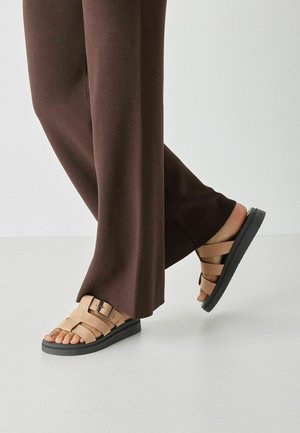 Tan leather sandals with black soles, featuring multiple wide straps and a side buckle, paired with brown ribbed pants.