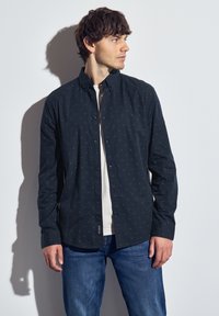 Námořnická košile s knoflíky a malými světlými vzory, dlouhými rukávy a límečkem, nošená přes bílý tričko, v kombinaci s modrými džíny.