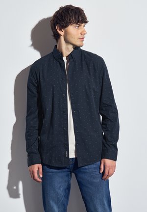 Camisa azul marino de botón con pequeños patrones claros, de mangas largas y cuello, llevada sobre una camiseta blanca, combinada con jeans azules.