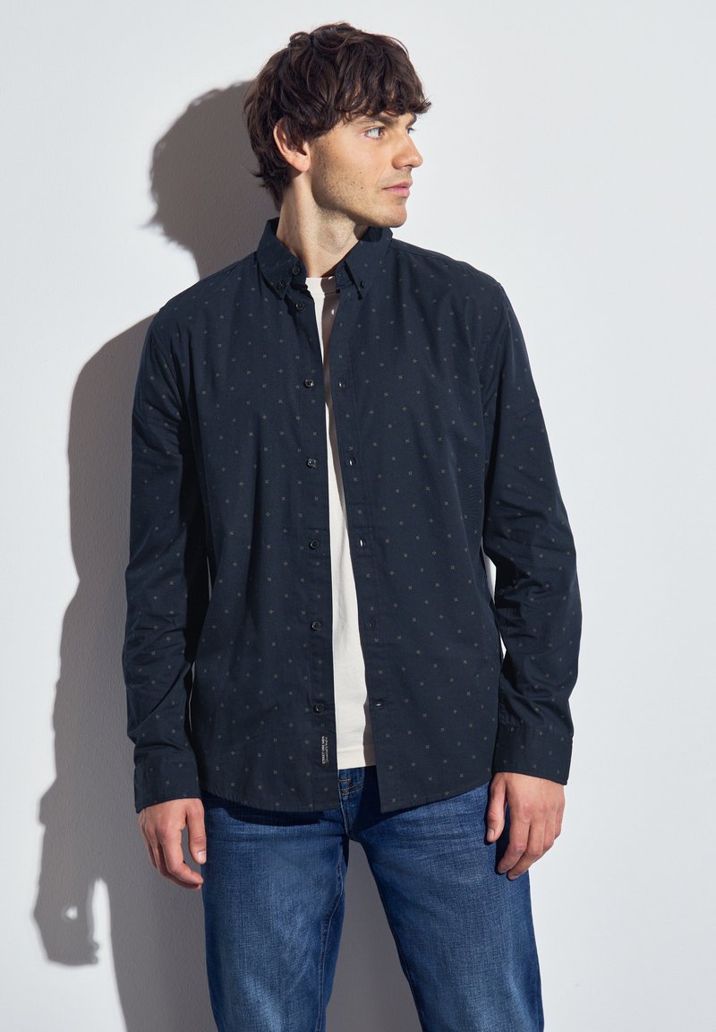 Námořnická košile s knoflíky a malými světlými vzory, dlouhými rukávy a límečkem, nošená přes bílý tričko, v kombinaci s modrými džíny.