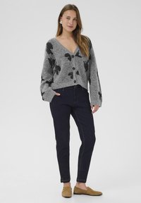 Grauer gestrickter Cardigan mit schwarzen Blattmustern, V-Ausschnitt und Knopfverschluss. Kombiniert mit dunklen Jeans und hellbraunen Slippern. Glatte Textur.