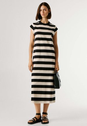 Frau trägt ein midi Kleid mit schwarzen und beigen horizontalen Streifen, hält eine schwarze Handtasche und trägt schwarze Sandalen. Sie steht vor einem einfarbigen Hintergrund.