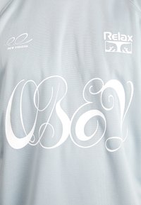 Světle modrý síťovaný svetr s výrazným bílým kurzivním textem „OBEY!“ a logo „New Visions“ a „Relax.“ Hladká textura, ležérní design.