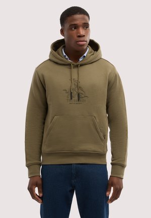 Junger Mann trägt olivgrünen Hoodie mit Fronttasche und Logo, kombiniert mit blauer Jeans, blickt leicht nach rechts vor einfarbigem Hintergrund.